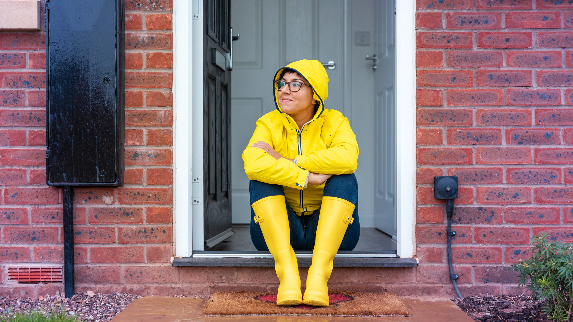 Frau mit Gummistiefeln und Regenjacke sitzt vor der Tür